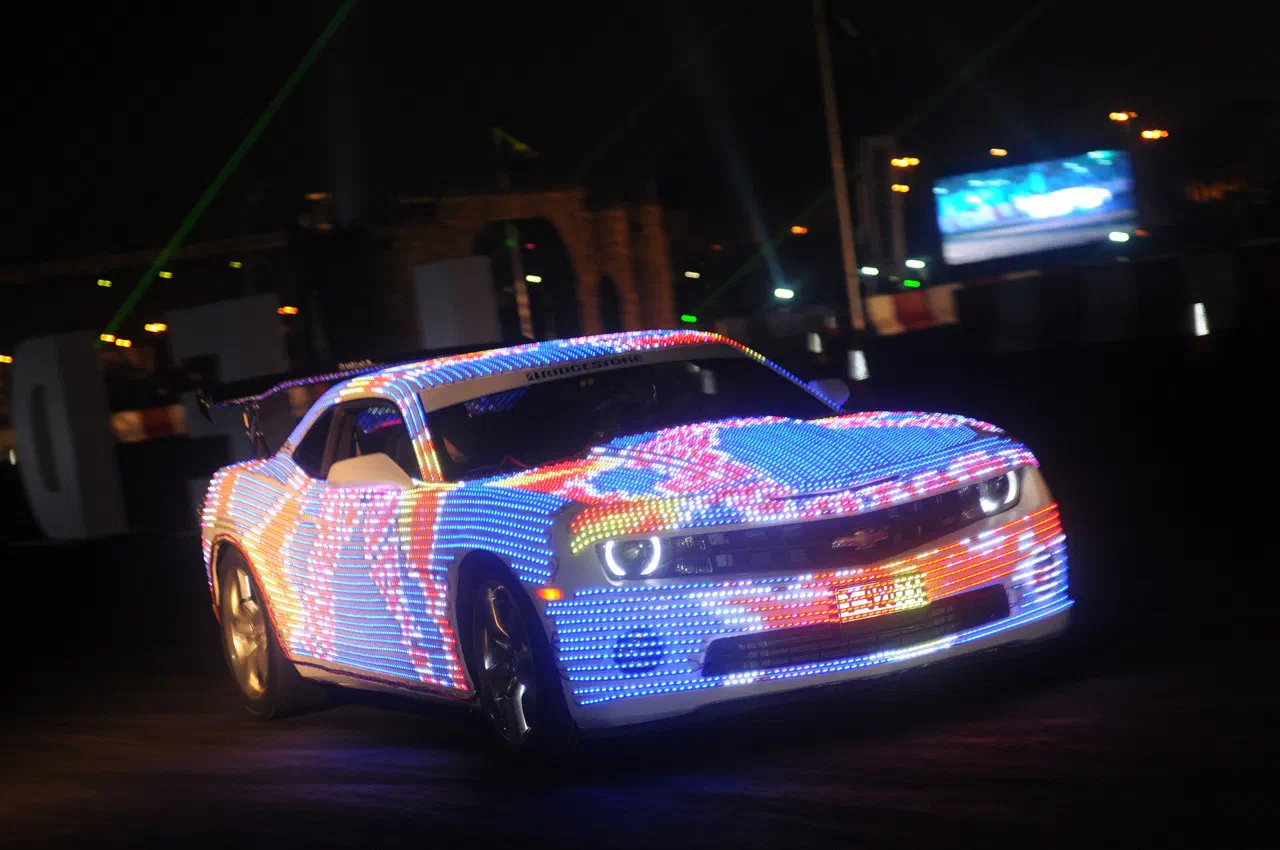 Chevrolet Camaro covered in colourful LED pixels driving at night, with green laser beams and a big screen in the background