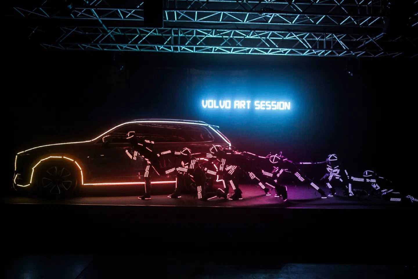 Tron dancers in glowing LED suits in a line pulling a Volvo SUV outlined in orange and pink LED light on a dark stage, under a neon “Volvo Art Session” sign