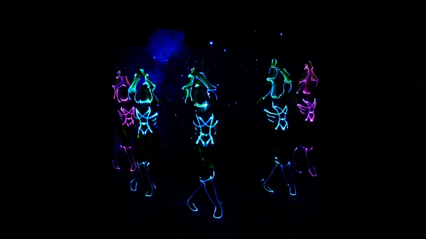 Five Tron Girls in blue, green and pink LED suits dance in front of an LED wall showing a blue galaxy