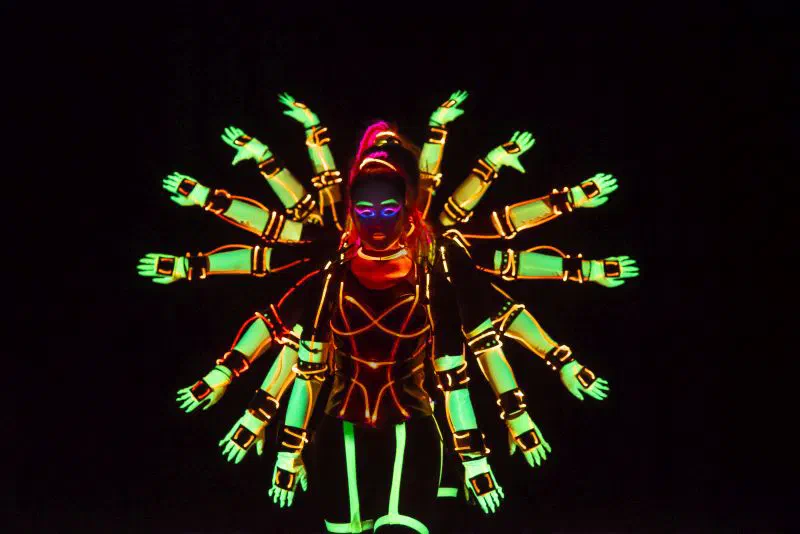 Tron Girls dancer in an orange LED and green UV costume with glowing face paint, the gloved arms of the dancers lined up behind her fanned out around her like a wheel