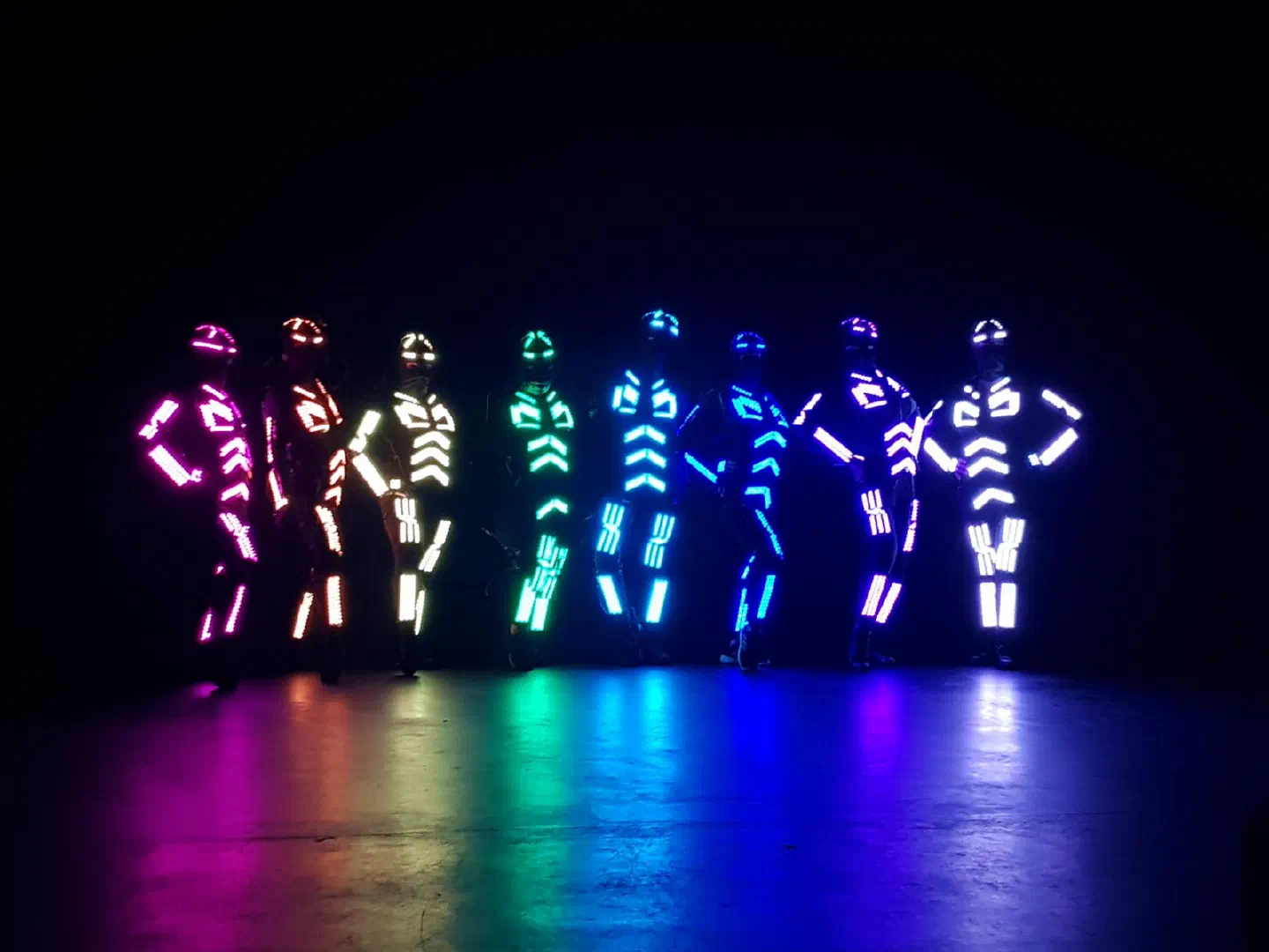 Eight dancers in our LED suits glowing pink, orange, white, green, cyan, blue, violet and lavender, posing in a row on a dark stage with coloured reflections on the floor