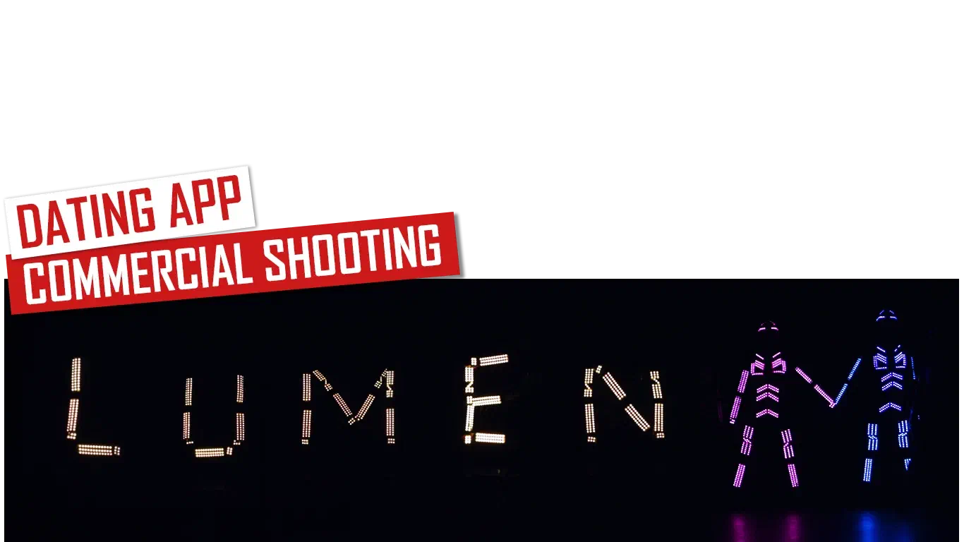 Dating app commercial shooting – red and white labels above the word LUMEN spelled in glowing LED letters, next to two dancers in pink and blue LED suits holding hands in the dark
