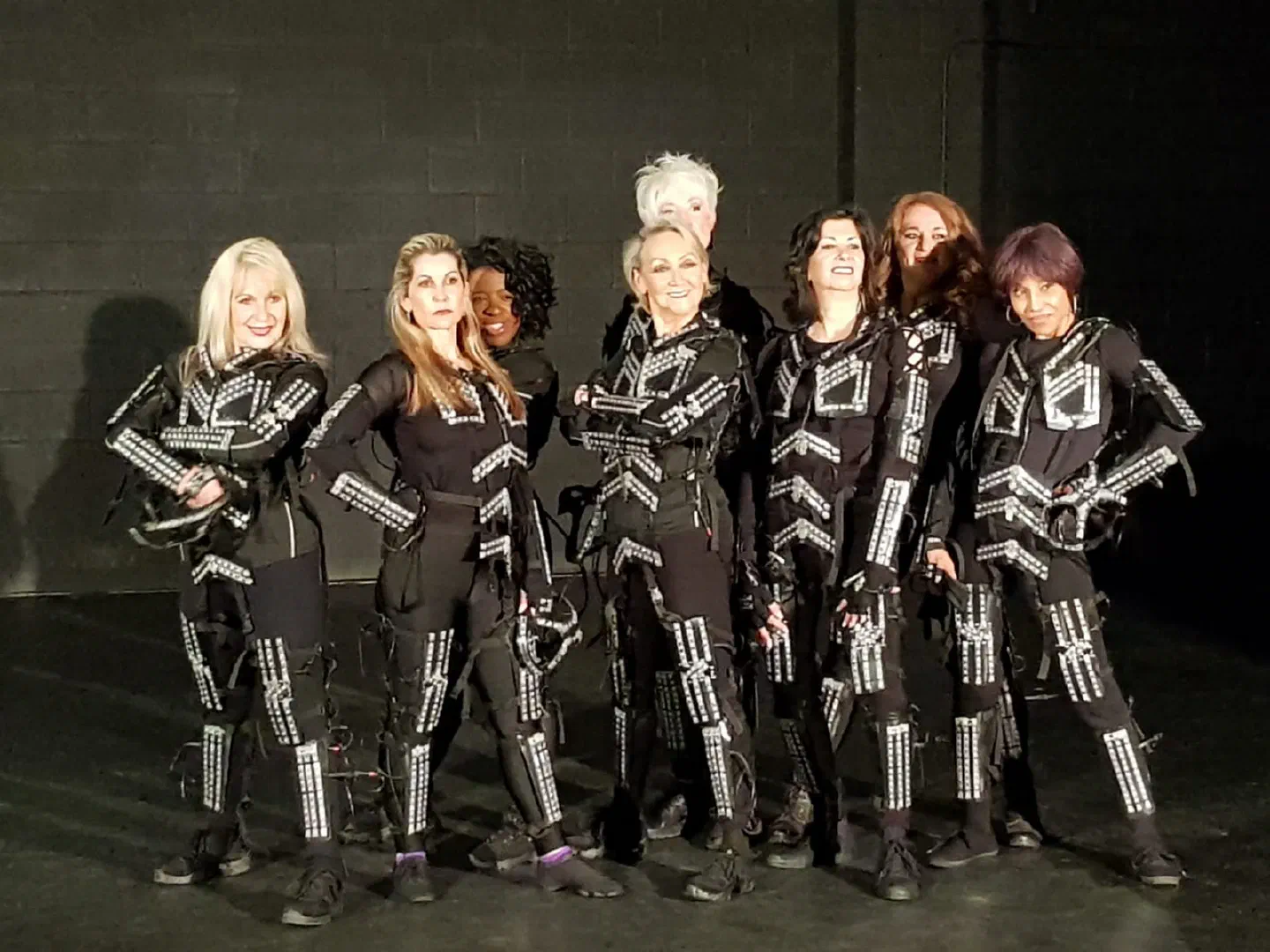 Eight ladies in black Tron Dance LED suits with the lights off, posing together on a dark studio floor in front of a black brick wall
