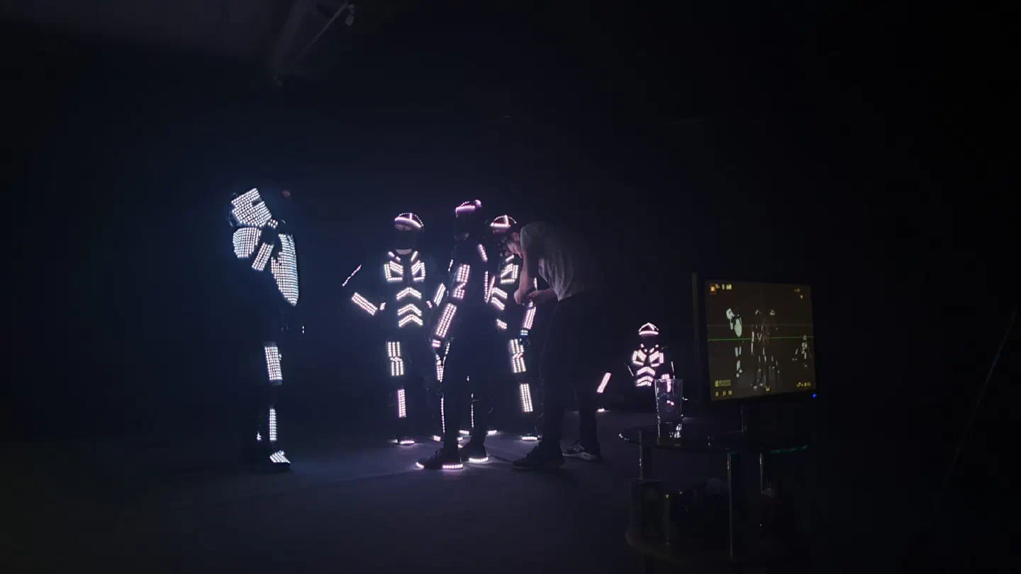 Dancers in glowing pixel and analog LED suits in a dark studio – a crew member adjusts one dancer’s suit, a monitor on the right shows the camera view of the dancers