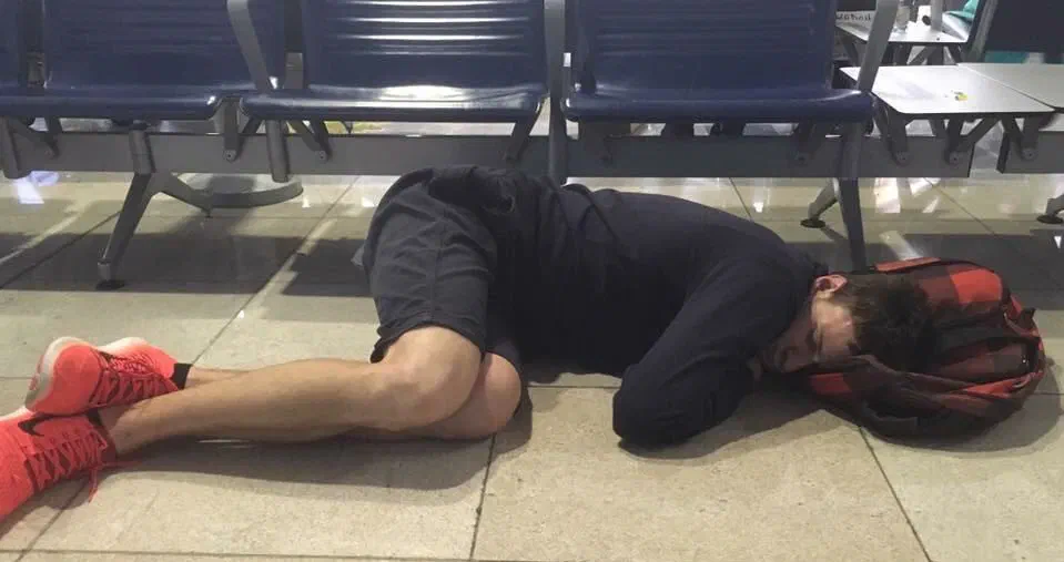 One of our team asleep on the floor of Dubai Airport, his head on a red checked backpack, under a row of blue seats