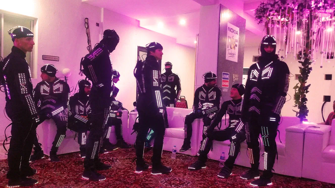 Tron dancers in black LED suits with glowing white LED strips waiting on white sofas in a pink-lit hall, with Nokia R&F and Leisure Leagues banners behind them