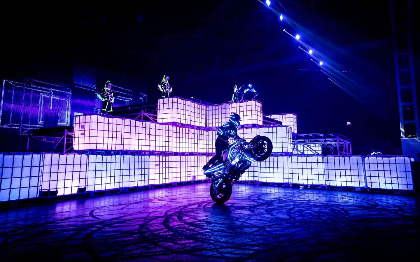 Jazdec v žiariacom LED kostýme robí na motorke wheelie pred stenou z fialovo osvetlených kociek, na ktorých tancujú Tron Girls v zelených LED kostýmoch