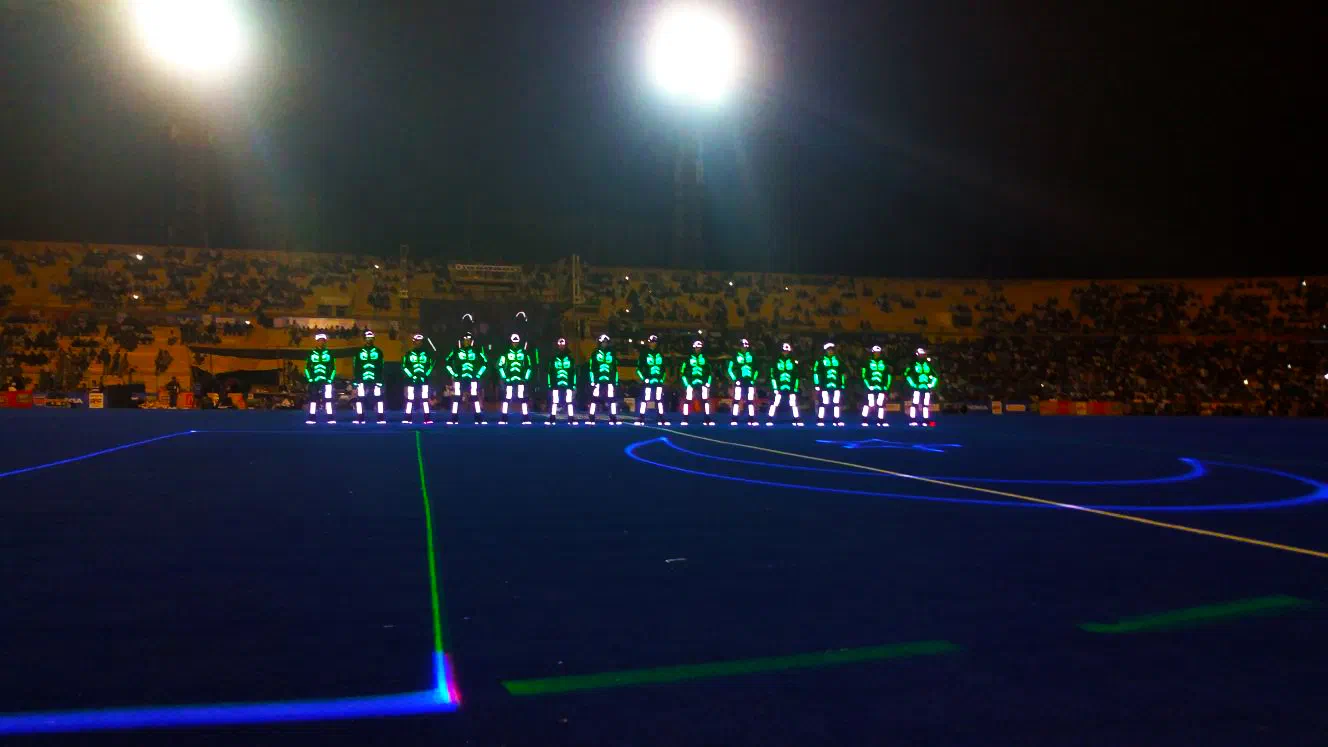 A row of Tron dancers in glowing green and white LED suits on the floodlit stadium pitch in Karachi, with laser lines drawn on the ground and spectators in the stands