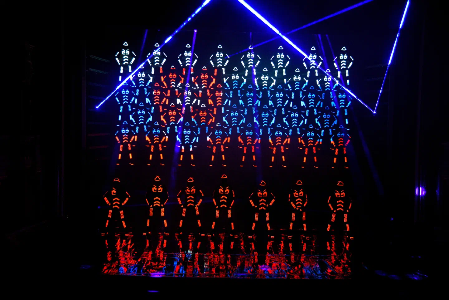 Tron dancers in red LED suits on stage in front of an LED screen that shows photos of the dancers in rows of white, blue and red – the Slovak flag – with blue laser beams above