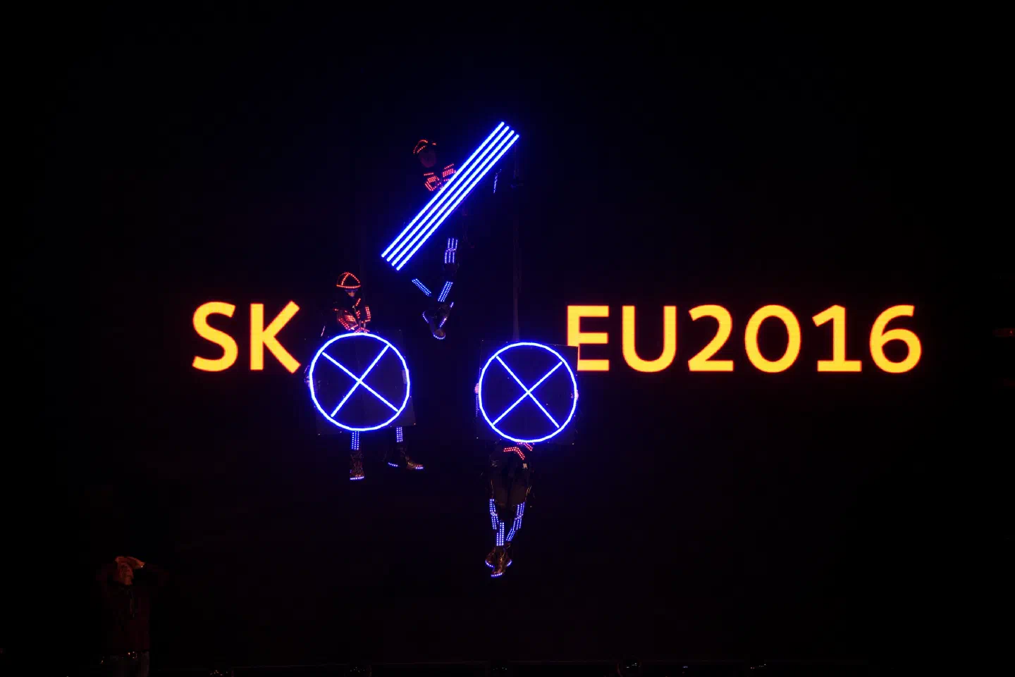 Three Tron dancers in LED suits hanging on ropes in the dark, holding the glowing blue parts of the Slovak presidency logo between the yellow letters SK and EU2016