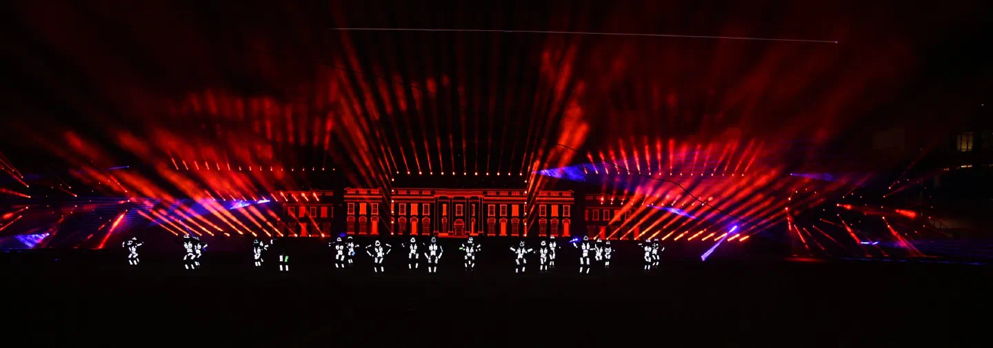 Tron dancers in white LED suits on the pitch of the Trnava football stadium at night, with red laser beams fanning out above them and a building facade glowing red behind