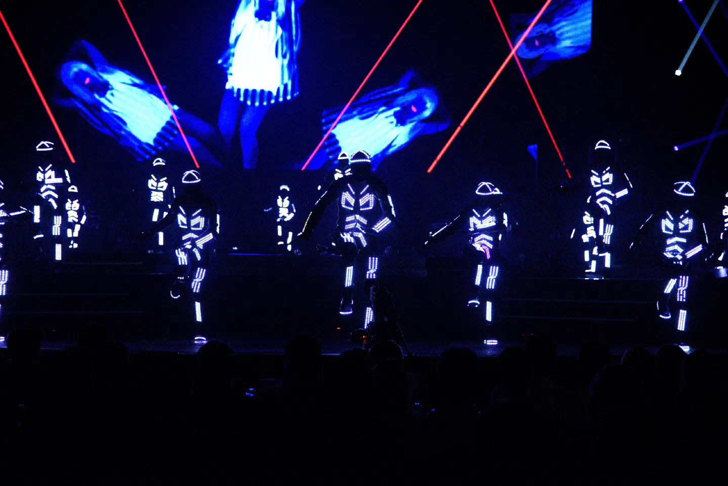 Tron dancers in glowing white LED suits on a dark stage at the Lucie Bílá show, with blue images on the LED screens and red laser beams behind them