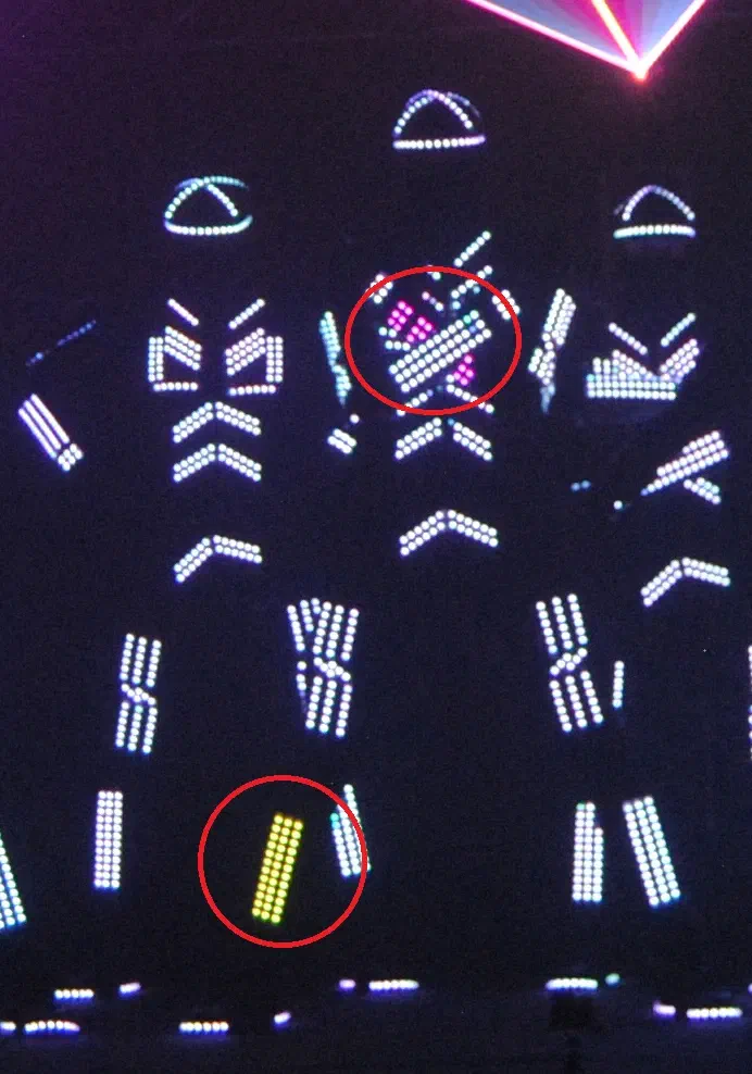 Three Tron dancers in LED suits glowing white on a dark stage – two suit parts circled in red show another colour: a pink and white chest part and a yellow leg part