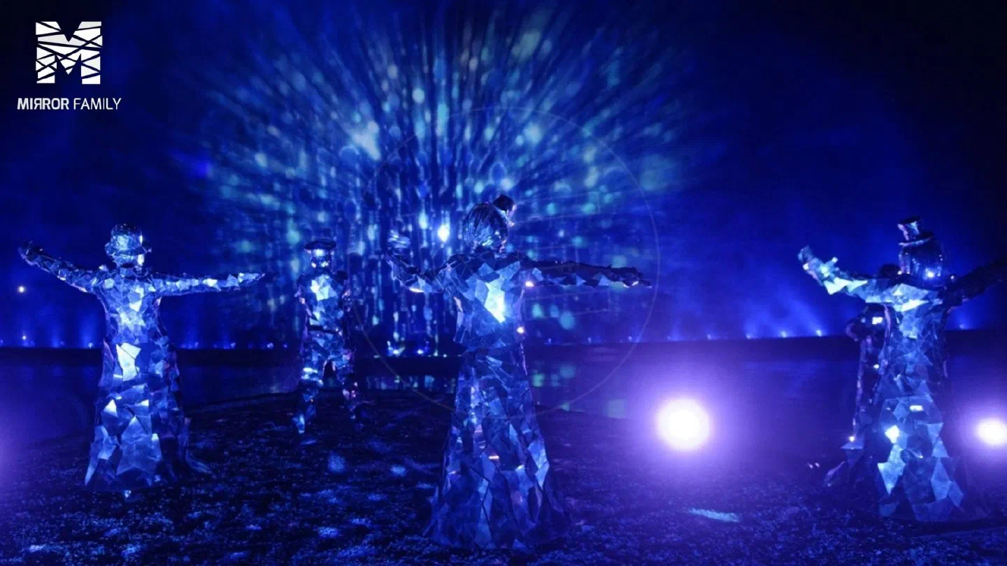 Five Mirror Family dancers in costumes covered with mirror shards, arms stretched out under blue light, with the Mirror Family logo in the corner