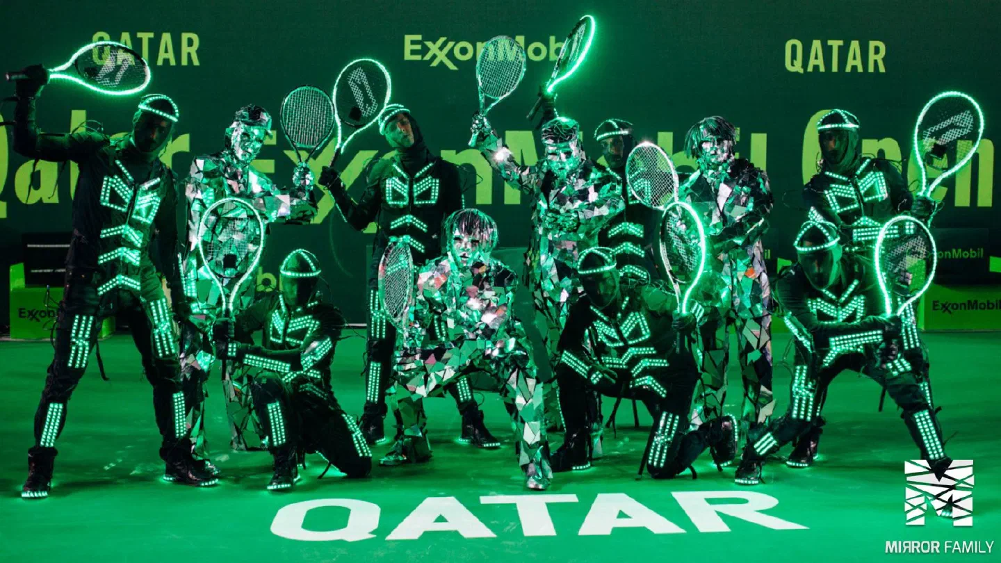 Tron dancers in green LED suits holding glowing LED tennis rackets, posing with Mirror Family dancers in mirror suits on a green court in front of Qatar ExxonMobil banners