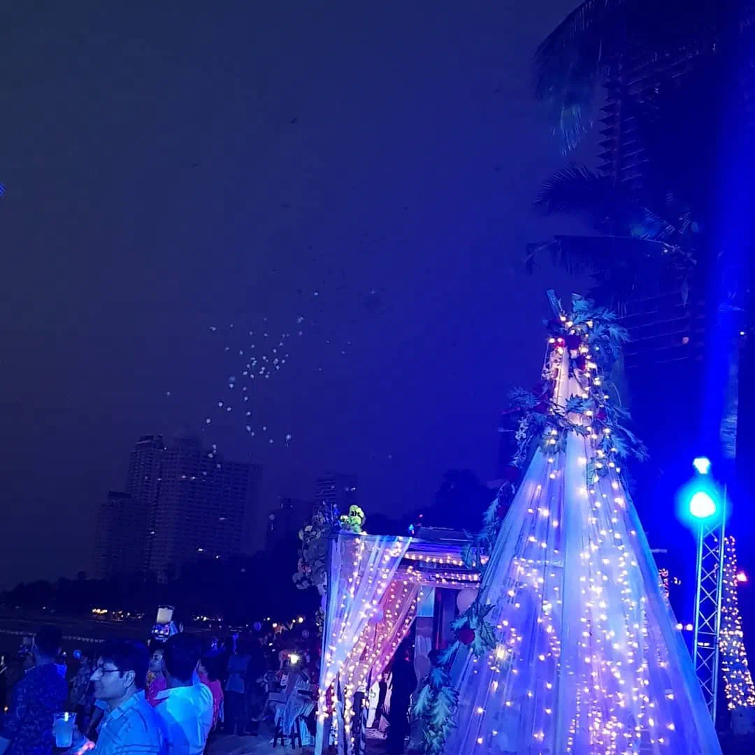 Wedding decoration on the beach at night – a tall cone and a canopy draped in white fairy lights and flowers, guests in front of them and lit balloons rising into the dark sky