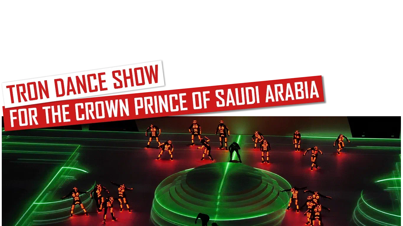 Show Tron Dance pre korunného princa Saudskej Arábie – červeno-biele banery nad tanečníkmi Tron v červených LED kostýmoch okolo okrúhleho kaskádového pódia, na podlahe zelené čiary futbalového ihriska