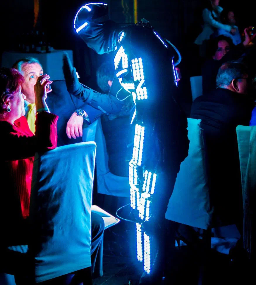 Tanečník Tron v čiernom kostýme so žiariacimi bielo-modrými LED pixelovými pásikmi a prilbou lemovanou LED sa nakláňa nad hostí pri stole, zatiaľ čo ho žena fotí mobilom