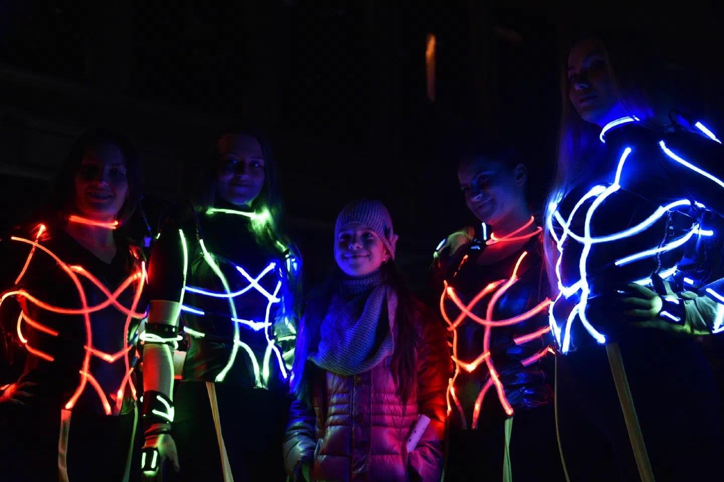 Štyri Tron Girls v LED kostýmoch s obrysmi žiariacimi na červeno, zeleno a modro pózujú v noci s usmiatym dievčaťom v ružovej bunde a pletenej čiapke, ktoré stojí medzi nimi