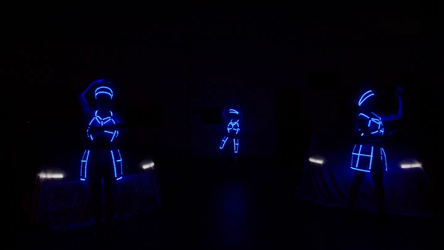 Tri tanečnice Tron Dolls v žiariacich modrých LED kostýmoch vystupujú v tme pred dvoma zakrytými autami