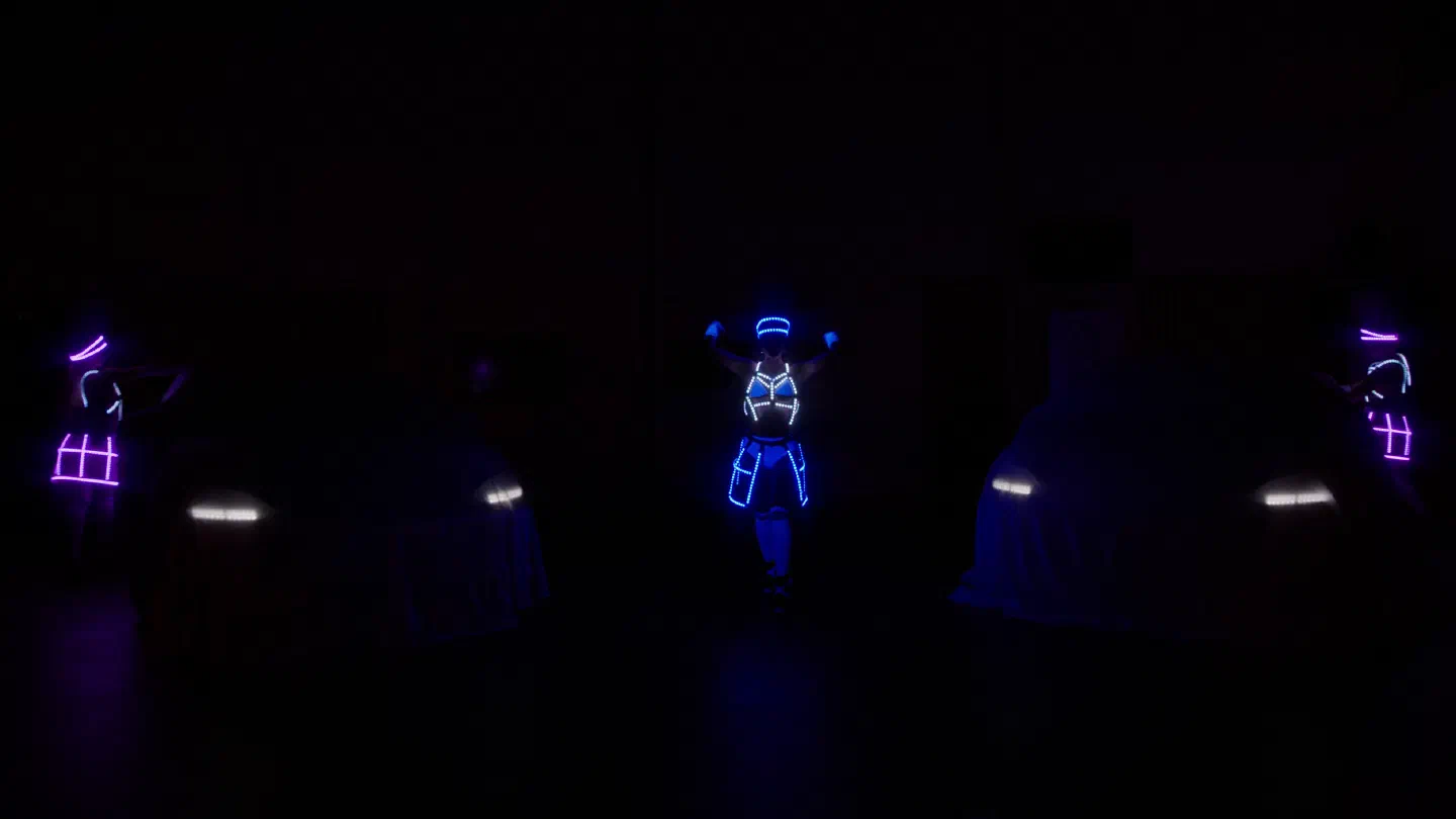 Tanečnice Tron Dolls v žiariacich modrých a fialových LED kostýmoch medzi dvoma zakrytými autami, ktorých svetlomety presvitajú cez plachty