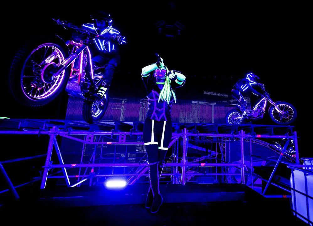Two FMX riders in LED-lit suits jumping their motorbikes on the stage behind a Tron Girl in a glowing costume with neon yellow braids, all in blue light