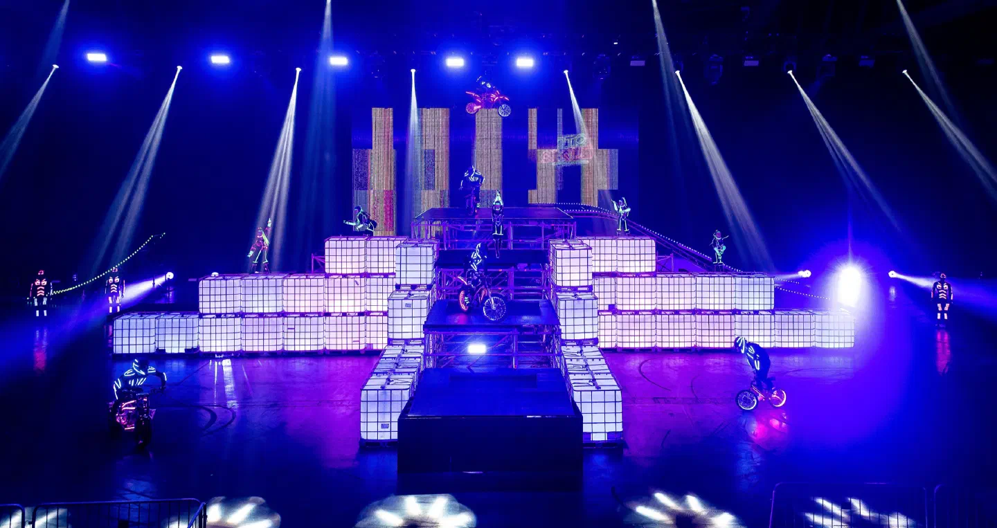 The Moto Cirque stage in a dark arena – a big construction of glowing white cube containers and steel platforms with an FMX rider in the air above it, riders and dancers in glowing costumes around it and white light beams from above