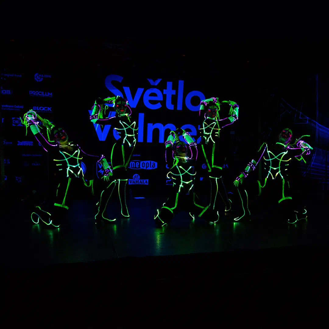 Five Tron Girls in LED & UV costumes glowing neon green and pink, posing on a dark stage in front of a blue Světlo Valmez banner with sponsor logos