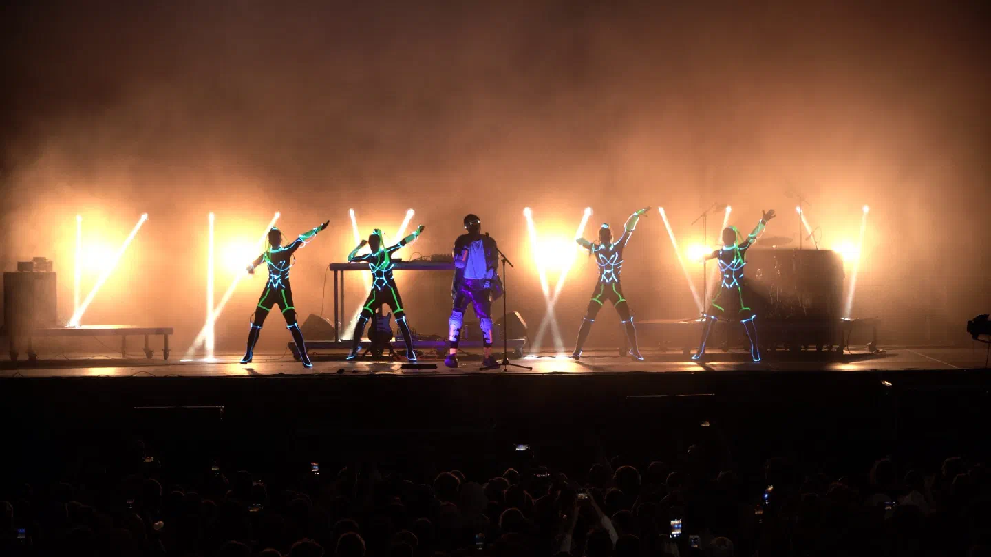 Four Tron Girls in glowing green and blue LED suits dancing around Fulcrum on the Pohoda main stage, backlit by white light beams in orange haze, with the crowd in front