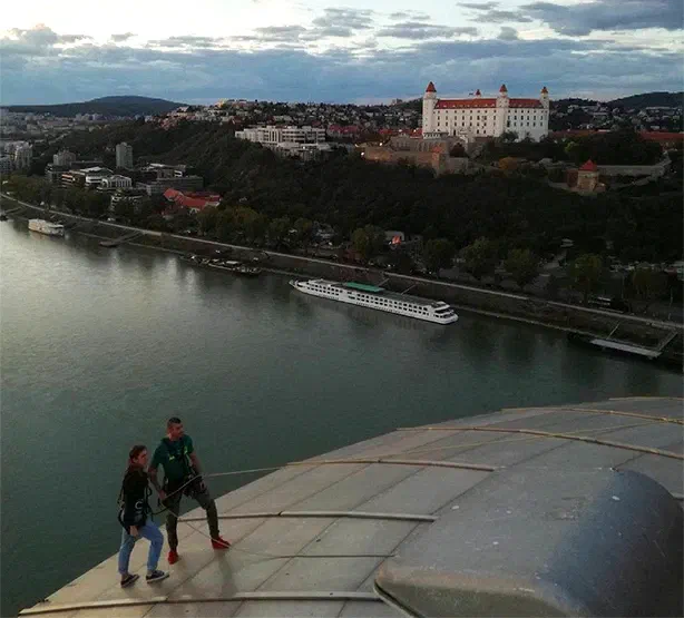 A dancer and a climber in climbing harnesses on the sloping roof of the UFO high above the Danube, with Bratislava Castle and a river cruise ship below