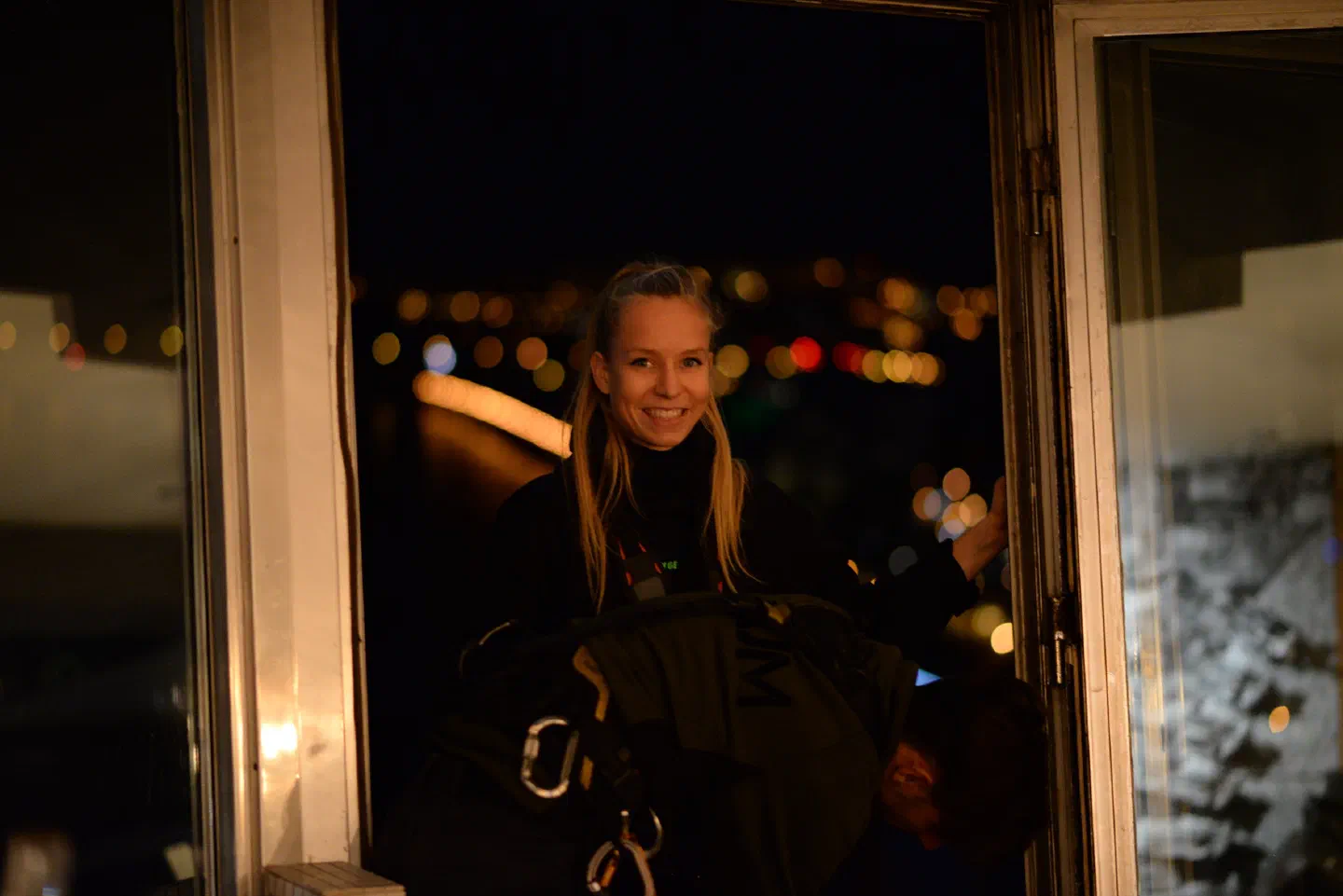 Tron Girl Veronika in a black outfit and climbing harness smiling in the open window of the UFO restaurant at night as a climber helps her out, with the city lights blurred behind her