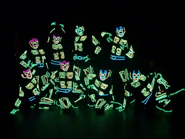 The Illuminate dance group – dancers in dark costumes outlined with glowing green, yellow and blue lines and glowing masks, posing together on a dark stage