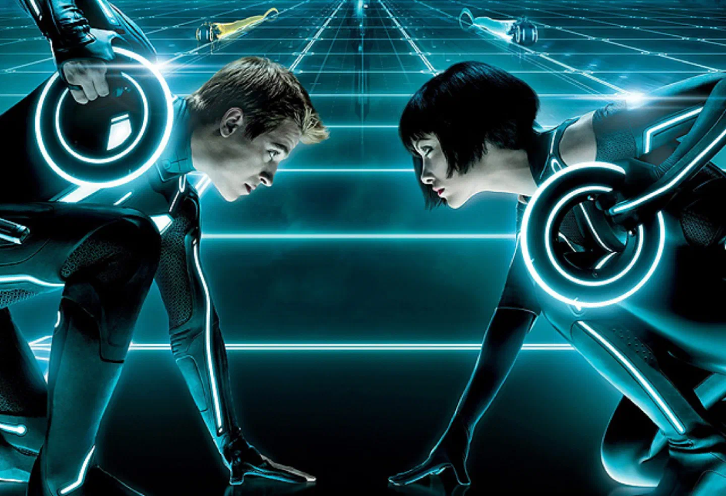 Two characters of the Tron Legacy movie in black suits with glowing turquoise lines and light discs on their backs, face to face on a glowing grid with light cycles behind them