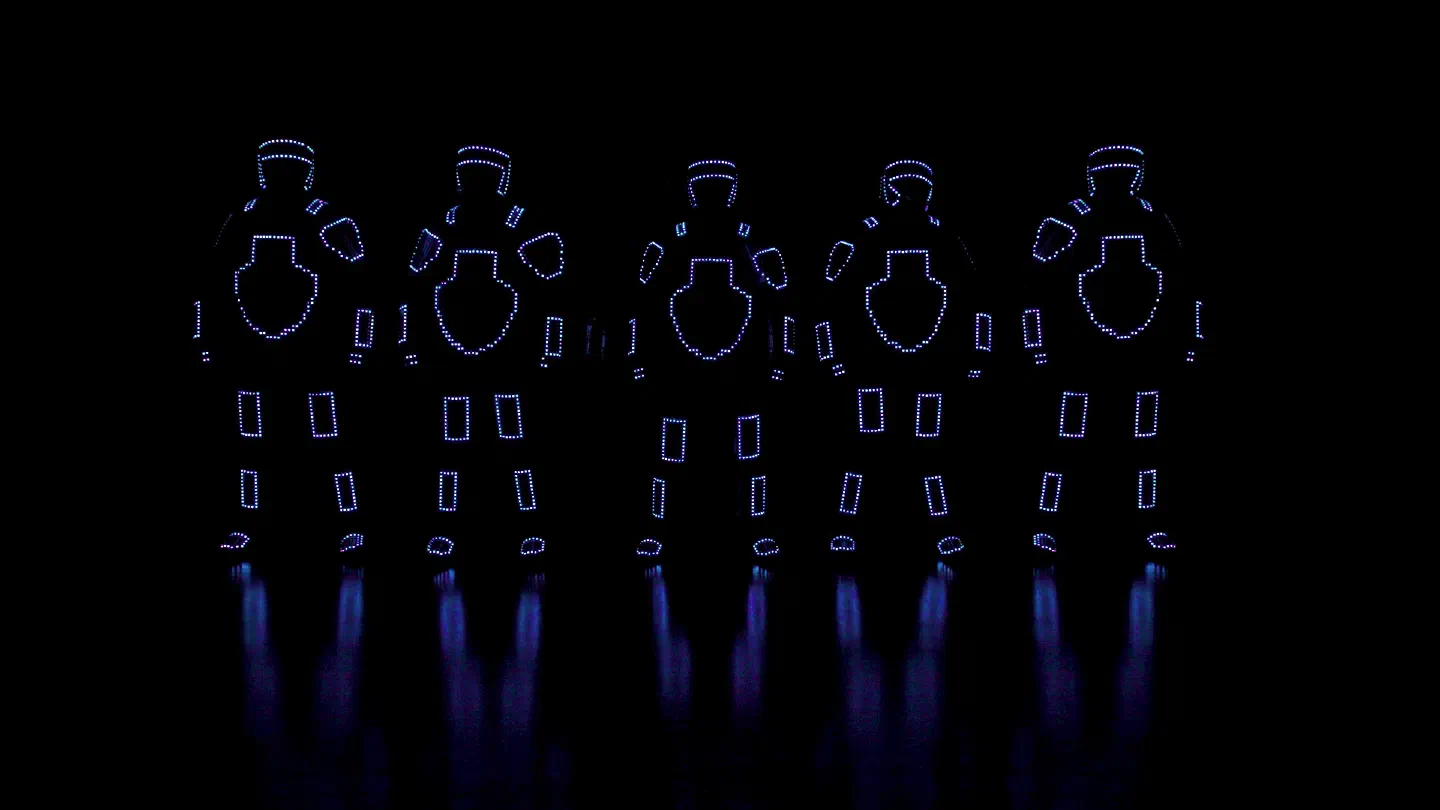 Five dancers in pixel LED suits standing in a row, only the outlines of their suits lit in dotted blue and violet pixels, reflected on the floor