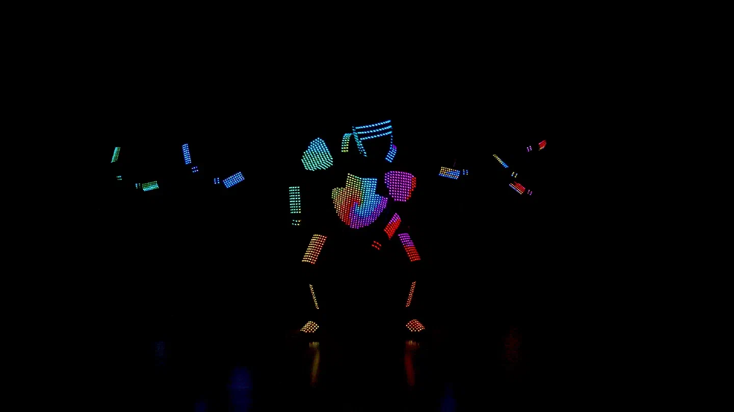 A dancer in a pixel LED suit glowing in rainbow colors in a wide stance on a dark stage, with the arms of two more pixel suits faintly lit on both sides
