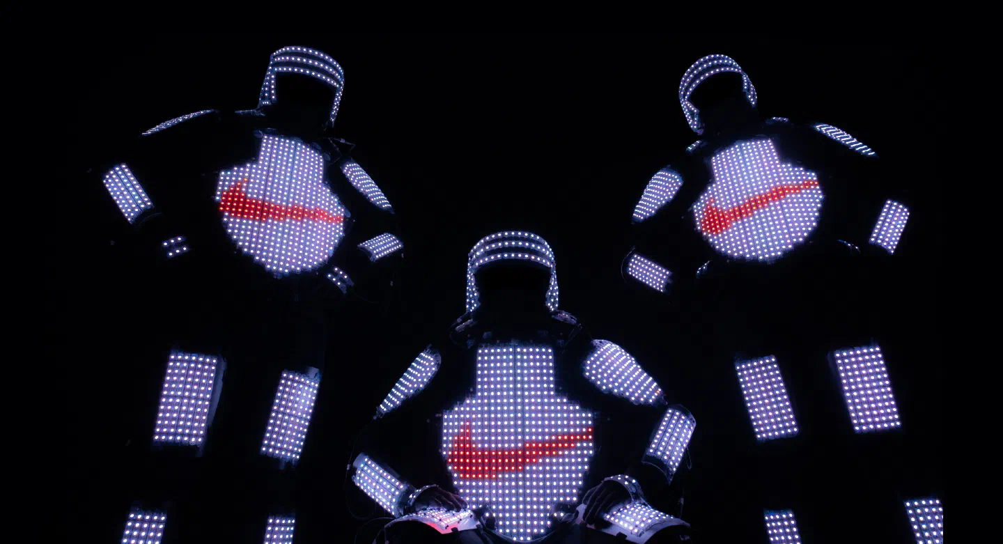 Three dancers in white LED pixel suits on a black background, each chest showing a red swoosh logo in pixels