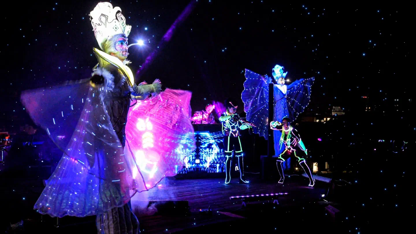 Performers in LED costumes on a rooftop stage at night – a stilt walker with a glowing crown and lit-up wings in front, a violinist and a dancer in Tron Girls LED suits and a DJ behind them, a second winged stilt walker on the right