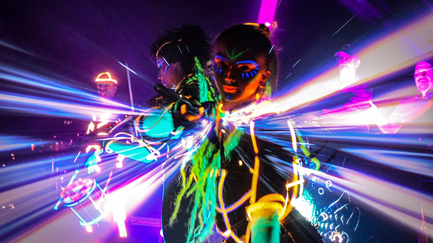 A violinist and a dancer with UV face paint in glowing orange and green LED costumes amid streaks of purple and blue light, an LED drummer with a lit-up hat on the left and the DJs on the right