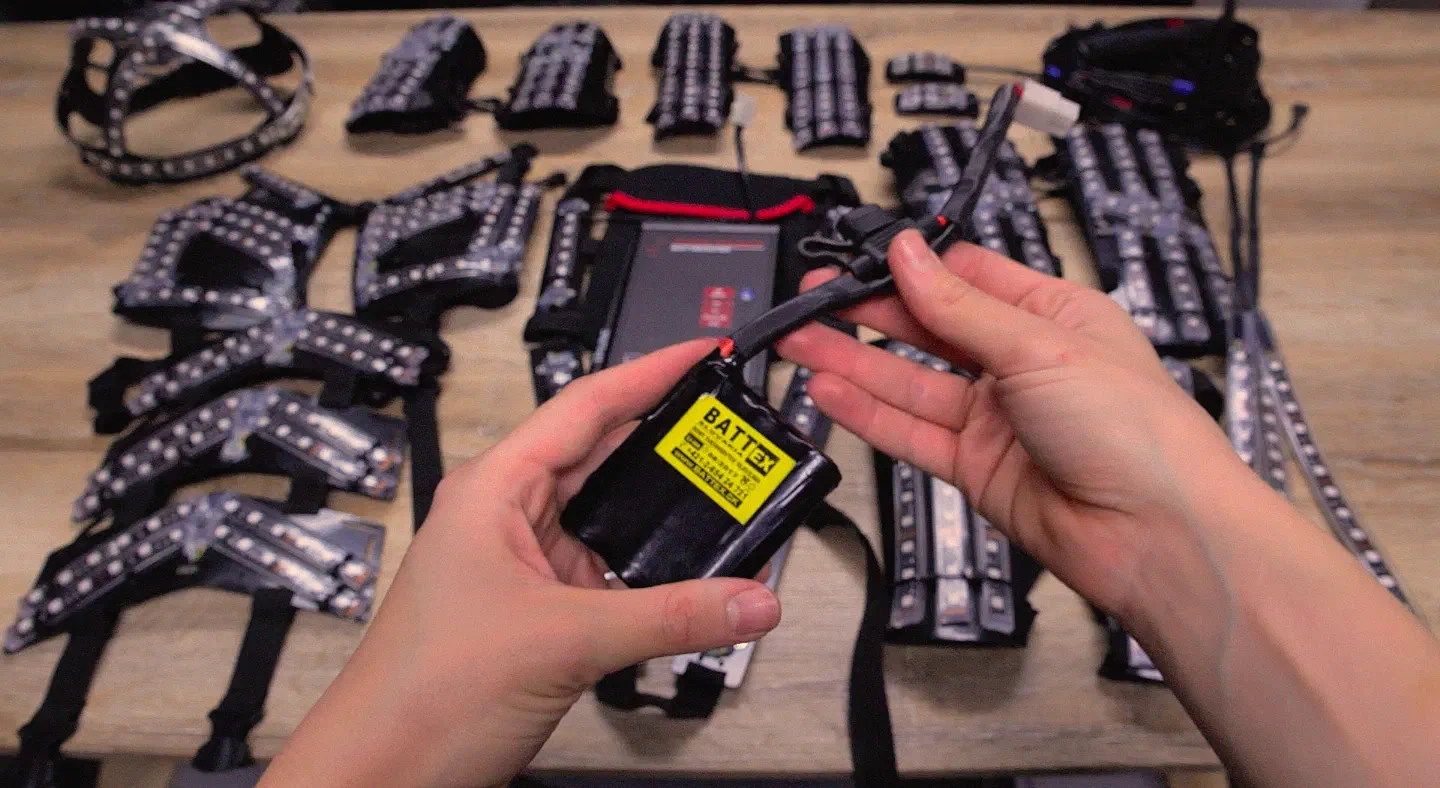Hands holding the small black Tron Dance battery pack with a yellow label and its cable, above the suit parts on a table