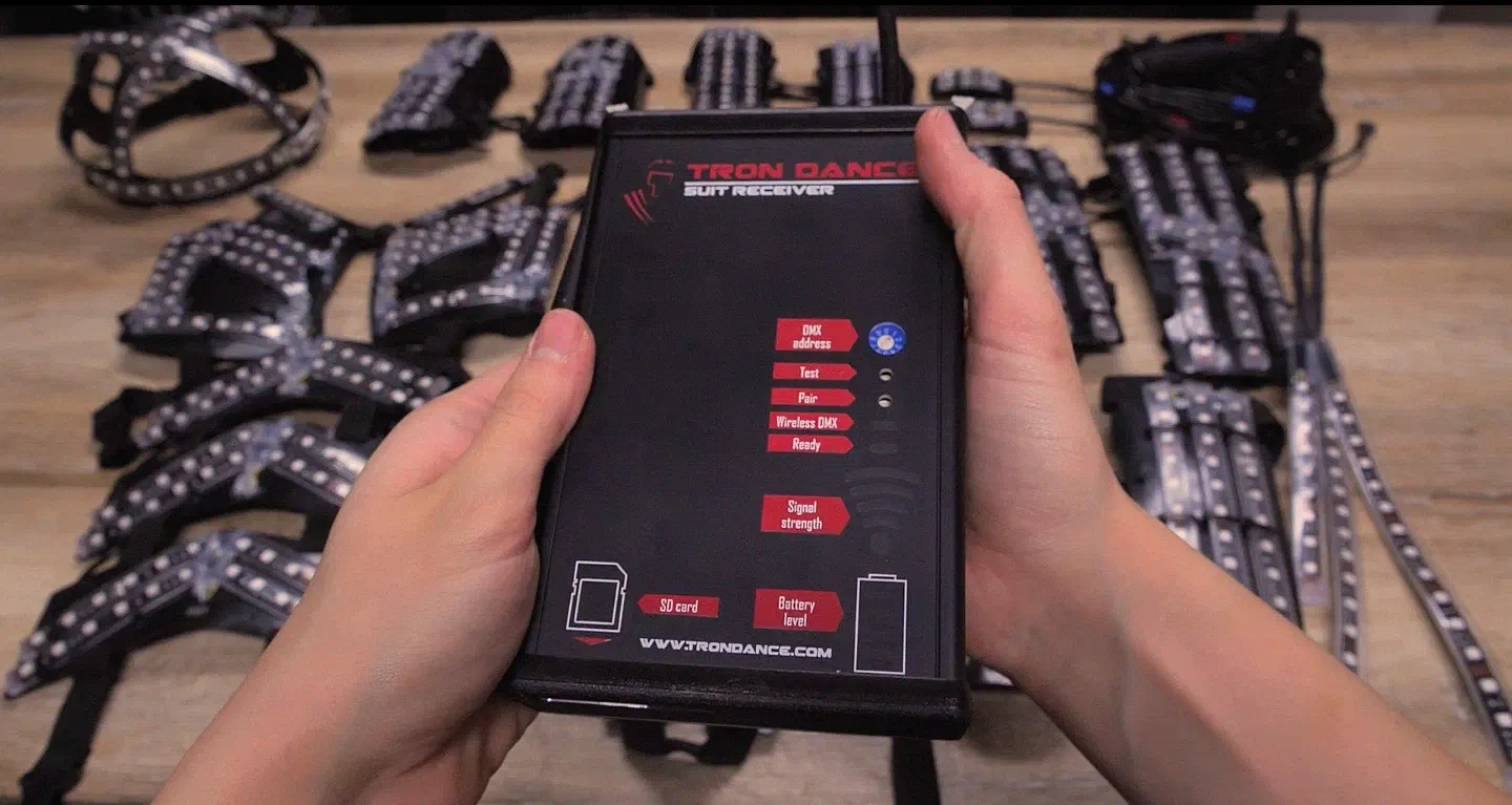 Hands holding the black Tron Dance Suit Receiver, with labels for DMX address, Test, Pair, Wireless DMX, Ready, Signal strength, SD card and Battery level, in front of the suit parts on a table