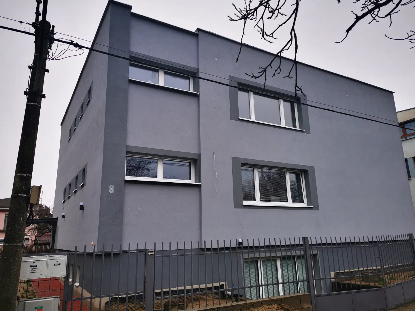 Nová budova našej spoločnosti – moderná sivá budova s bielo orámovanými oknami za tmavým kovovým plotom v zamračený deň