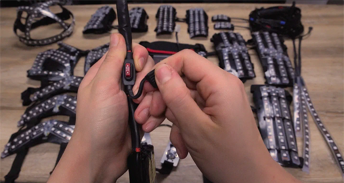 Hands opening the fuse holder on the battery cable to show the red 10 A fuse, with LED suit parts on a wooden table behind