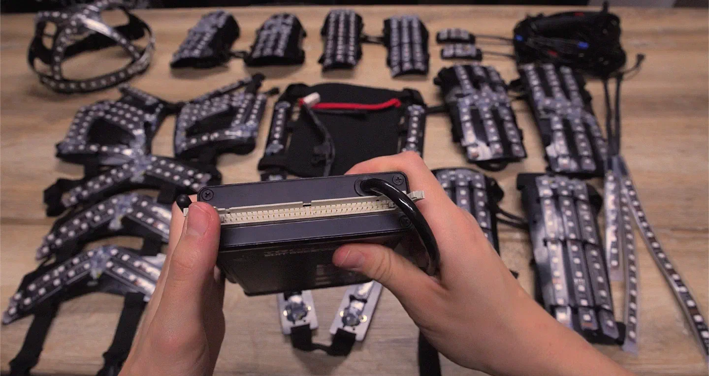 Hands holding a Tron Dance suit receiver upside down to show the row of pins in the empty connector on its top edge, next to the black cable leading out of it, with LED suit parts on a wooden table behind