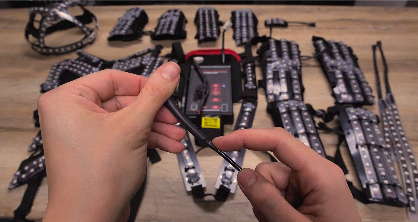 Hands holding the thin cable of one LED suit part in front of the receiver on the back plate of a Tron Dance suit, with the other suit parts laid out on a wooden table
