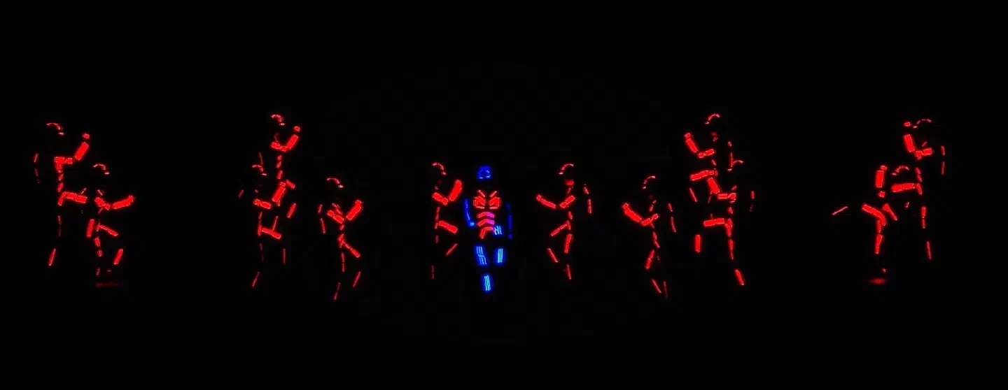 The same LED dance crew with their suits glowing red around one dancer in a blue LED suit in the middle of the dark stage