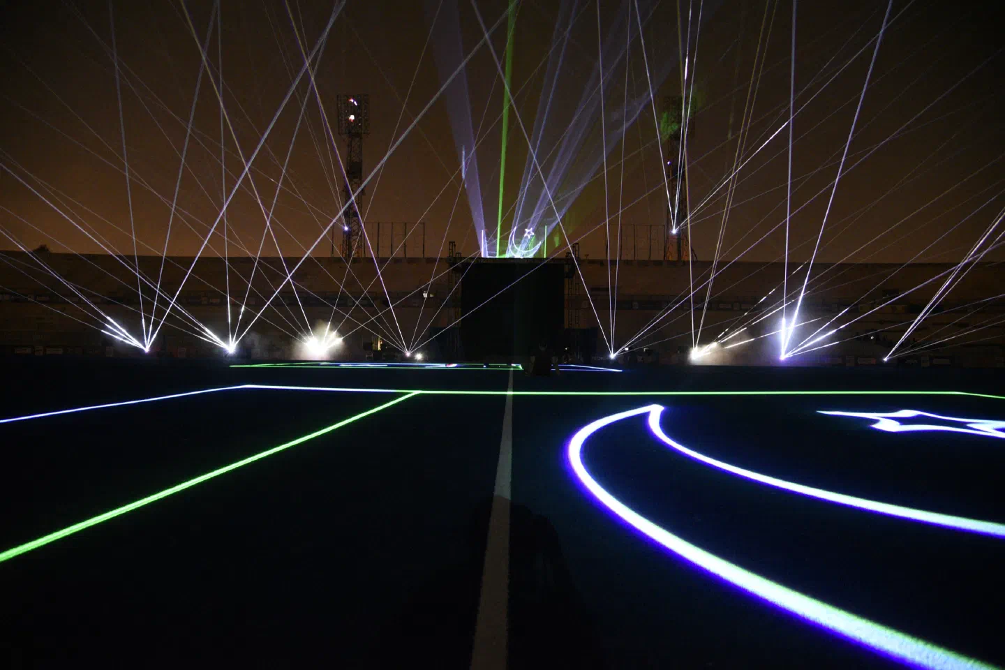 White and green laser beams fanning out over the stadium in Pakistan at night, with laser lines, a crescent moon and a star drawn on the dark pitch