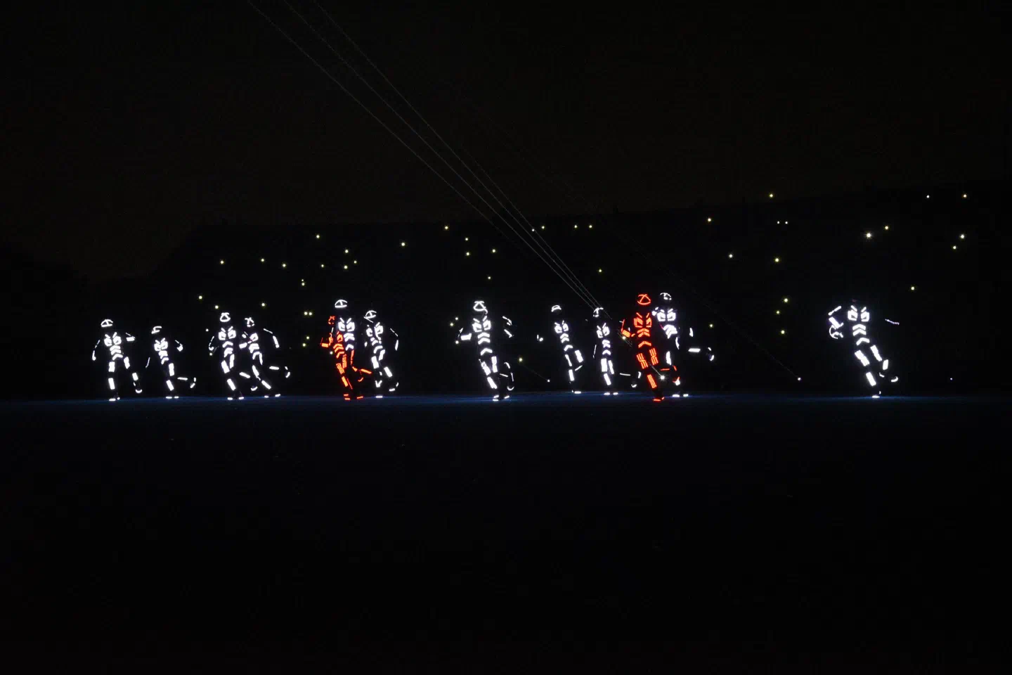 Tron dancers in LED suits glowing white, two of them in red, dancing on the dark stadium pitch in Pakistan, with laser beams above them and small lights in the stands