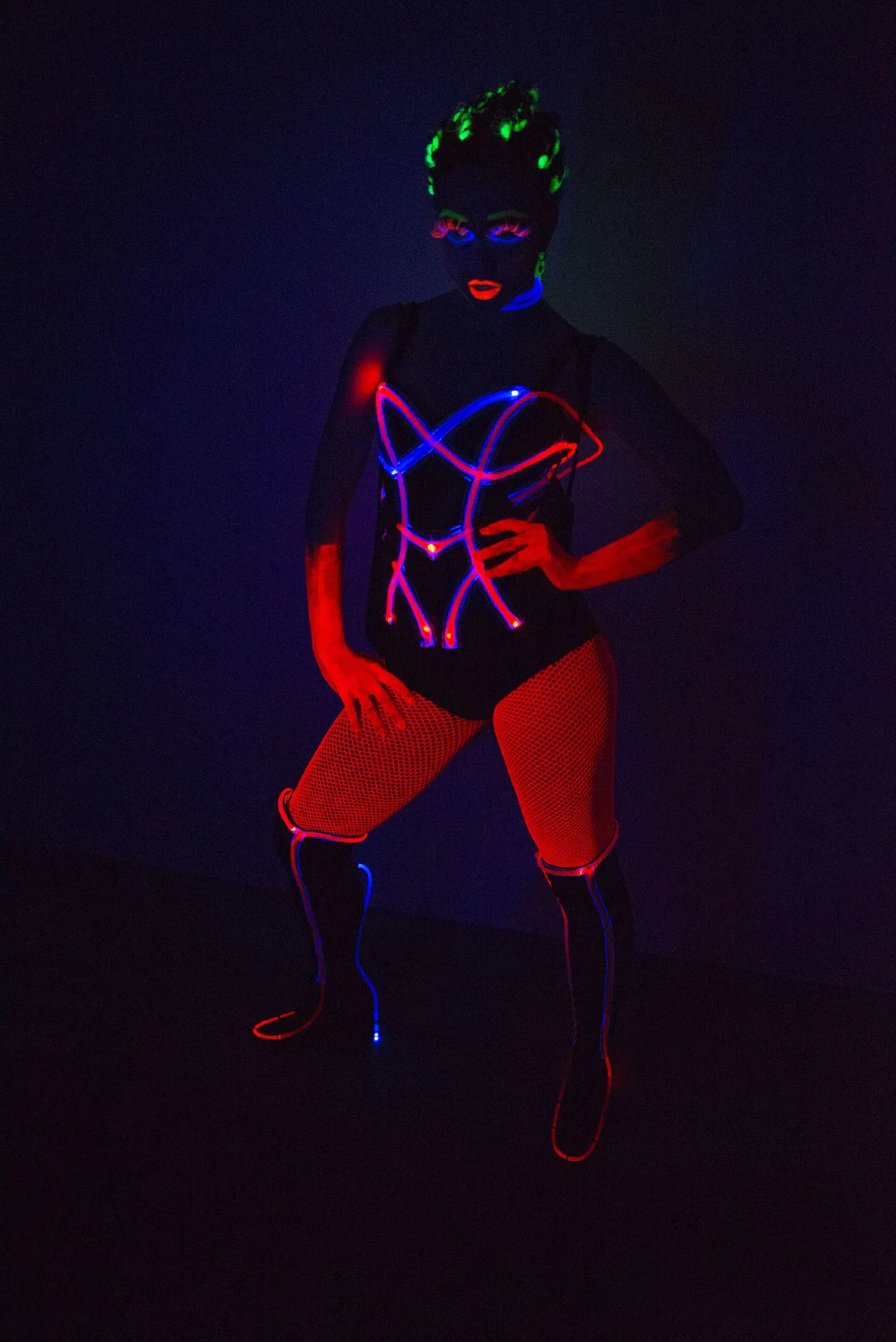 Tron Girls dancer in a black bodysuit and fishnet tights, with glowing red and blue lines across the bodysuit and the boots, UV make-up and fluorescent green hair, posing in the dark