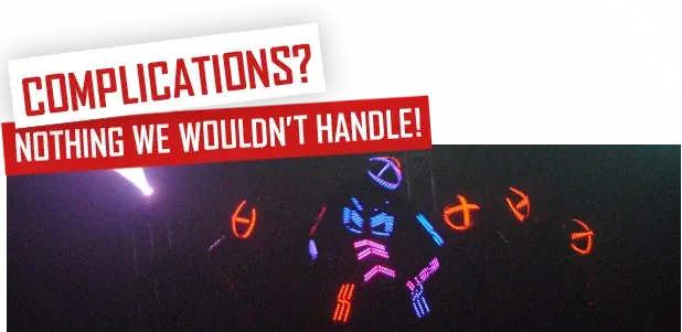 Complications? Nothing we wouldn’t handle! – white and red labels above dancers in glowing multicoloured LED suits on a dark stage, with a laser beam on the left