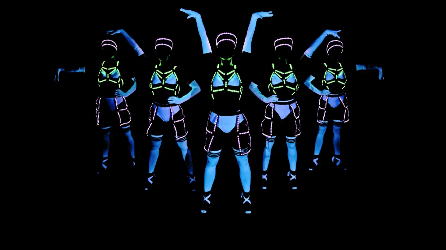 Five Tron Girls in white costumes glowing blue under UV light, posing with raised arms, their LED harnesses glowing green on the chest and pink on the caps and hips
